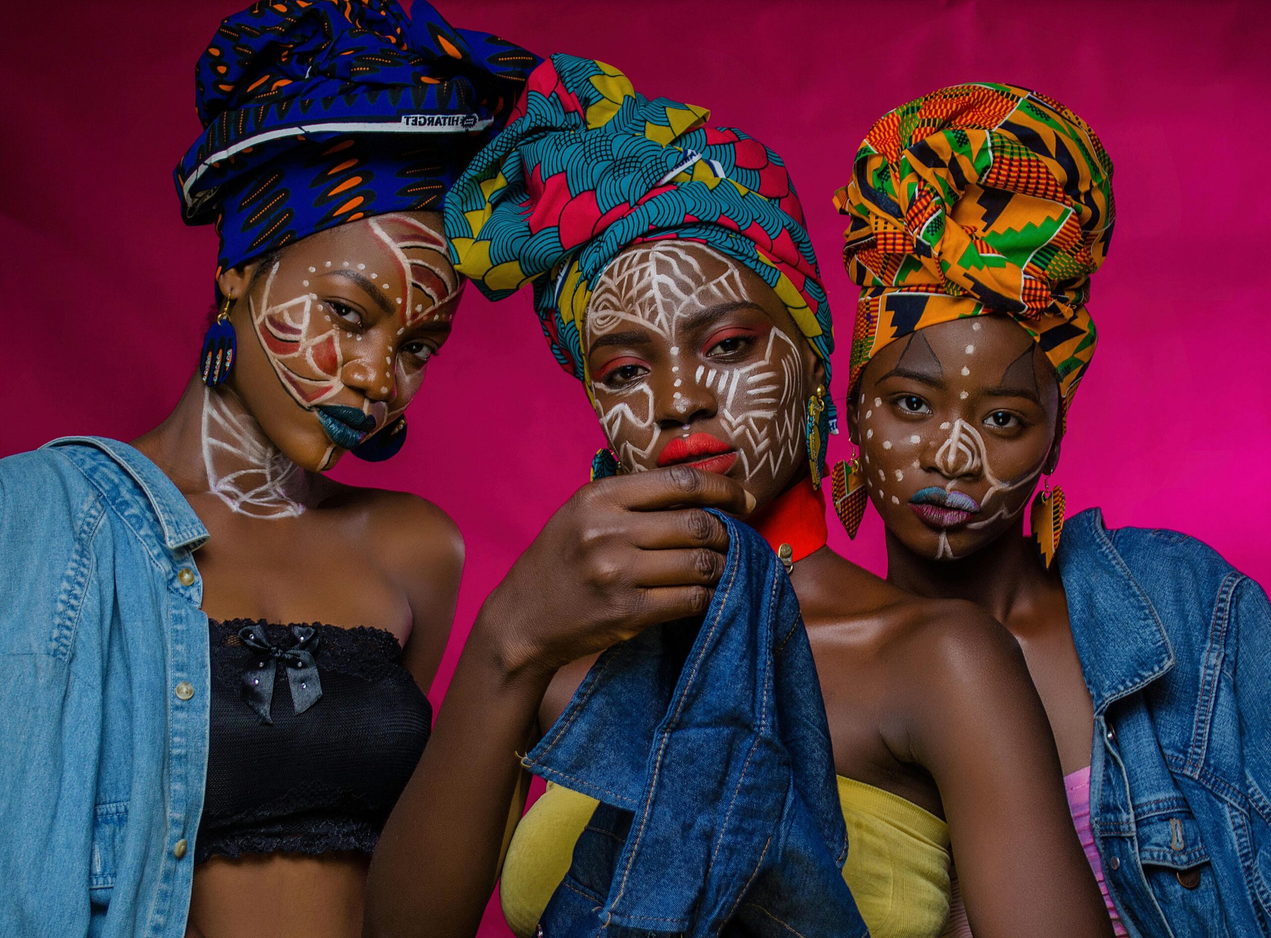 Striking portrait of three women showcasing African fashion with colorful headwraps and intricate face paint.